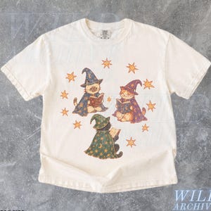May include: An ivory short-sleeve t-shirt featuring a graphic print of three cats wearing wizard hats and capes, each reading a book. The cats are surrounded by orange star illustrations. The shirt has a crew neckline and a standard fit.