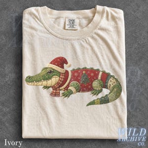 May include: An ivory t-shirt featuring a graphic of a green alligator wearing a red and white Santa hat, a plaid scarf, and a red jumper with a Christmas tree pattern. The alligator has a quilted texture design and the shirt has a Comfort Colors label.