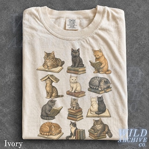 May include: An ivory crew neck t-shirt featuring a graphic print of twelve cats in various poses, many interacting with stacks of books. The shirt has a Comfort Colors brand label at the inner neckline and is made of cotton fabric.