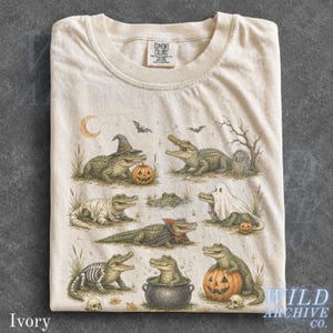 May include: An ivory t-shirt featuring a graphic print of various alligators in Halloween costumes, including a witch, a ghost, a vampire, and a skeleton. The design includes pumpkins, a tombstone, bats, and a crescent moon on a cotton fabric.