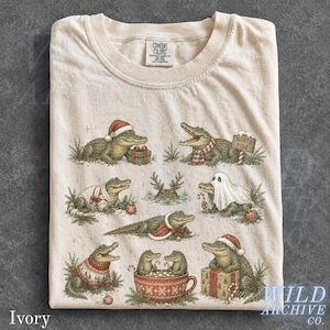 May include: An ivory t-shirt featuring a graphic print of various alligators in holiday attire. The illustrations include alligators wearing Santa hats, scarves, and sweaters, sitting in a mug, or holding gifts, with a North Pole sign included.