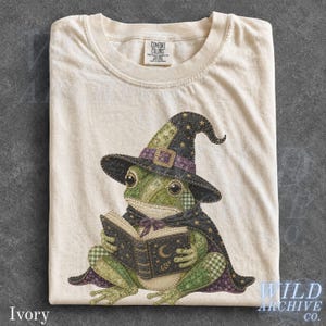 Op de afbeelding: Een ivoorkleurig t-shirt met een afbeelding van een groene kikker die een donkerpaarse en zwarte heksenhoed en cape draagt, terwijl deze een open boek met een halve maan vasthoudt. Het Comfort Colors label is zichtbaar in de nek.