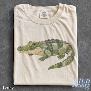 May include: An ivory t-shirt featuring a graphic of an alligator composed of various green and cream-coloured patchwork patterns. The shirt has a crew neckline and a Comfort Colors brand label printed on the inside of the collar area.