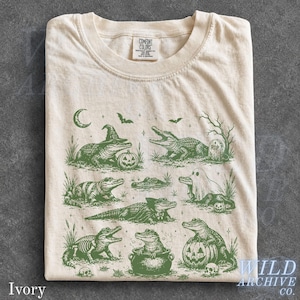 May include: An ivory t-shirt featuring a dark green graphic print of various alligators in Halloween-themed costumes and settings, including a witch, a ghost, a vampire, and a skeleton. The Comfort Colors brand label is visible at the neck.