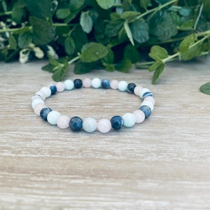 May include: A beaded bracelet featuring a repeating pattern of smooth, round beads in shades of pale pink, light blue, and dark navy blue. The beads are strung together on an elastic cord, forming a circular shape with a polished, uniform finish.