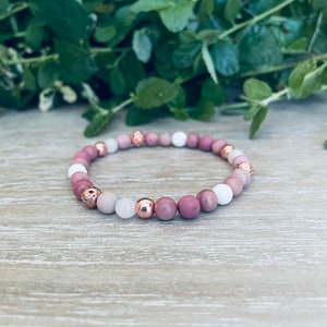 Puede incluir: Una pulsera de cuentas con un patrón repetitivo de cuentas redondas de piedra rosa, cuentas blancas lisas y cuentas metálicas de acento en tono oro rosa. El diseño incluye cuentas esféricas pulidas y cuentas metálicas porosas y texturizadas.