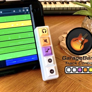 Könnte beinhalten: Eine weiße Makro-Tastatur mit fünf vertikalen Tasten, die Symbole für Kopfhörer, Stummschaltung, Automatisierung sowie Pfeile nach oben und unten zeigen. Die Tasten sind gelb, orange, lila und weiß. Das Gerät ist als GarageBand Track Control beschriftet.