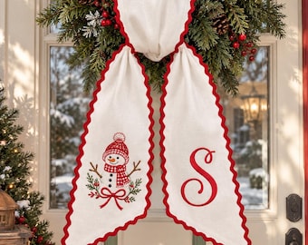 May include: A white fabric door hanger featuring a red scalloped edge. One side displays an embroidered snowman wearing a red hat and scarf, while the other side features a large, red embroidered letter S in a cursive script font.