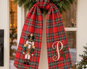 May include: A red, green, and yellow plaid fabric door hanger featuring an embroidered nutcracker on the left side and an embroidered letter P with a small holly berry accent on the right side, hanging from a green wreath.
