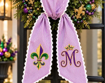 May include: A purple and white striped fabric door hanger with scalloped edges. The left side features an embroidered gold, purple, and green fleur-de-lis. The right side features an embroidered purple letter M topped with a gold crown.