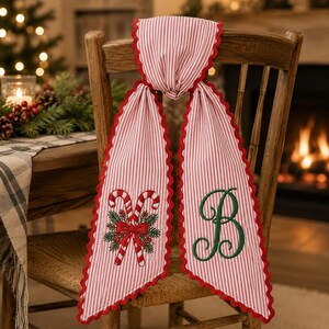 May include: A red and white striped fabric chair sash featuring a scalloped red edge. One side displays an embroidered design of two candy canes with pine needles and a red bow, while the other side features a large, dark green embroidered letter B.