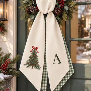 Peut inclure: Un accessoire de porte en tissu crème orné d'un sapin de Noël brodé en vert avec un nœud rouge d'un côté et une lettre A vert foncé de l'autre. L'envers du tissu présente un motif à carreaux vichy vert et blanc.