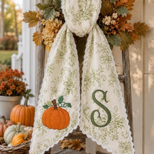 May include: A cream-colored fabric banner with a green floral print and white scalloped edges. The left side features an embroidered orange pumpkin with green leaves, while the right side displays a large, dark green embroidered letter S.