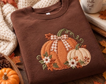 May include: A dark brown crewneck sweatshirt featuring an embroidered design of two pumpkins, one orange and one cream, accented with a white and brown gingham bow, small white flowers, and green leaves, with a ribbed collar and cuffs.