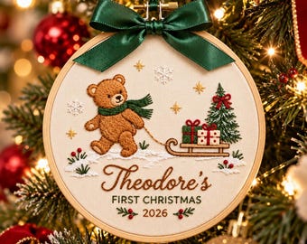May include: A round embroidery hoop ornament featuring a brown teddy bear pulling a wooden sled with gifts and a Christmas tree. The fabric is cream-colored with embroidered snowflakes and the text "Theodore's FIRST CHRISTMAS 2026". A dark green satin ribbon bow is attached to the top.