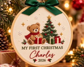 May include: A round wooden embroidery hoop ornament featuring a tan teddy bear holding a red gift, a green Christmas tree, and two small gifts. The text reads MY FIRST CHRISTMAS Charles 2026. A dark green satin ribbon bow is attached at the top.