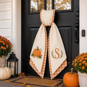 May include: A cream-colored fabric door hanger featuring an orange gingham border. One side displays an embroidered orange pumpkin with green leaves, while the other side features an embroidered orange letter S framed by two green leafy branches.