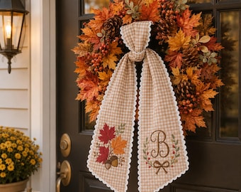May include: A fall wreath featuring orange, red, and yellow maple leaves, pinecones, and red berries. A beige and white gingham ribbon hangs from the center, embroidered with a red maple leaf, acorns, and a letter B surrounded by a wreath.