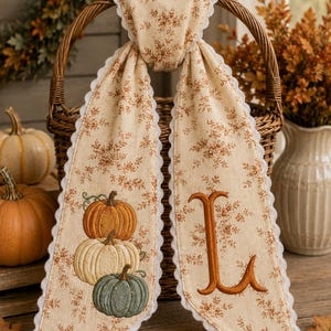 May include: A beige fabric ribbon with a brown floral pattern and white scalloped edges, draped over a wicker basket handle. One side features an embroidered stack of three pumpkins in orange, cream, and green, while the other side displays a large letter L.