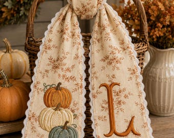 May include: A beige fabric ribbon with a brown floral pattern and white scalloped edges, draped over a wicker basket handle. One side features an embroidered stack of three pumpkins in orange, cream, and green, while the other side displays a large letter L.