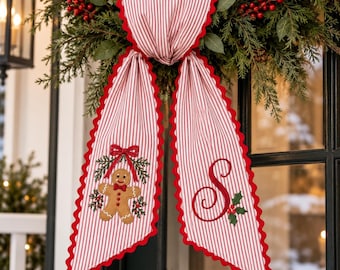 May include: A red and white striped fabric door hanger with scalloped edges. One side features an embroidered gingerbread man with a red bow, and the other side displays a large red embroidered letter S with holly leaves and red berries.
