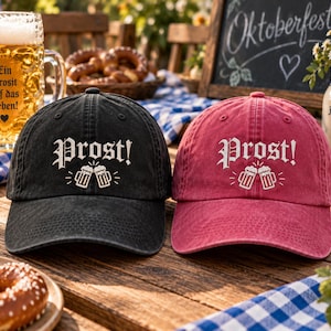 May include: A black baseball cap and a red baseball cap, both featuring the white embroidered text Prost! above a graphic of two clinking beer mugs. Each cap has a structured crown, a curved brim, and a washed, vintage-style fabric finish.