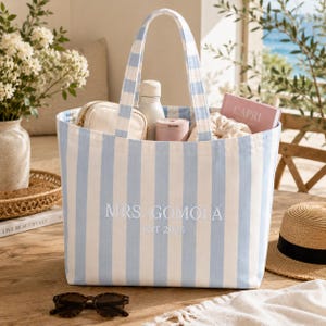 May include: A light blue and white vertical striped canvas tote bag featuring the embroidered white text MRS. GOMOLA EST 2026 on the front. The bag has two matching striped shoulder straps and is filled with various personal items.