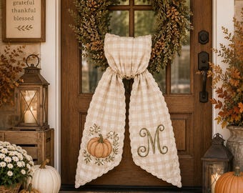 May include: A beige and white gingham fabric door hanger featuring a scalloped edge, an embroidered orange pumpkin on the left tail, and a large embroidered letter M on the right tail, suspended from a green foliage wreath on a wooden door.