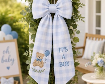 May include: A large decorative bow with a light blue and white gingham pattern and a 50-inch sash. One tail features an embroidered brown teddy bear holding a blue balloon, while the other tail displays the embroidered text IT'S A BOY.