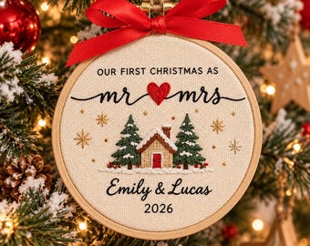 May include: A round wooden embroidery hoop ornament featuring cream fabric with embroidered text reading OUR FIRST CHRISTMAS AS mr mrs, a red heart, a cabin, two pine trees, and the names Emily & Lucas with the year 2026. A red ribbon bow is tied at the top.