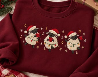 May include: A dark red crewneck sweatshirt featuring an embroidered design of three cream-colored sheep wearing red Santa hats. Each sheep holds a red mug and is wrapped in a string of colorful Christmas lights, surrounded by musical notes and snowflakes.