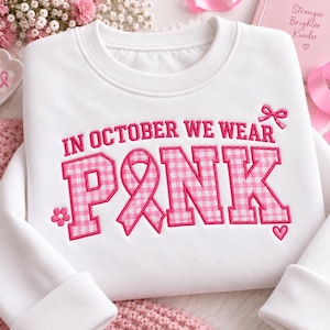 Op de afbeelding: Een wit sweatshirt met ronde hals en de geborduurde tekst IN OCTOBER WE WEAR PINK. Het woord PINK is ontworpen met een roze en wit ruitpatroon en de letter I is vervangen door een roze bewustwordingslint.