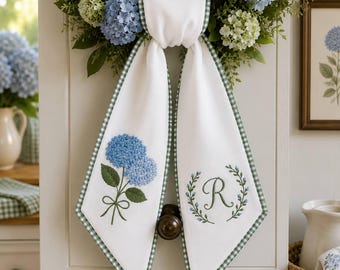 May include: A white fabric door hanger featuring a green gingham border. The left panel displays an embroidered blue hydrangea flower with green leaves, while the right panel features an embroidered letter R surrounded by a green leaf wreath.