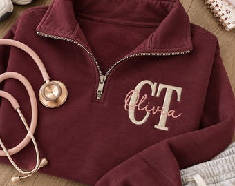 May include: A maroon quarter-zip sweatshirt featuring an embroidered monogram with the letters CT in a large cream font and the name Olivia in a smaller pink script font. A light pink stethoscope is positioned on the left side of the garment.