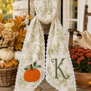 May include: A cream-colored fabric scarf featuring a green floral pattern, white scalloped trim, an embroidered orange pumpkin with green leaves on one end, and a large green embroidered letter K on the other end, draped over a black metal frame.