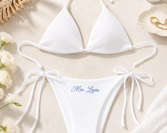 May include: A white two-piece bikini set featuring a triangle top with halter neck ties and matching bikini bottoms with side ties. The bikini bottoms are embroidered with the blue text Mrs. Levin in a cursive script font.