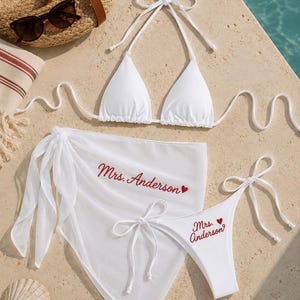 May include: A white bikini set consisting of a triangle top and side-tie bottoms, paired with a matching sheer sarong. The sarong and bikini bottoms feature the embroidered red text Mrs. Anderson with a small red heart icon.