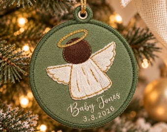 May include: A round, olive green embroidered ornament featuring a white angel with brown hair and a gold halo. The text "Baby Jones" and "3.8.2026" is stitched in white below the angel. A gold cord is attached to the top loop.