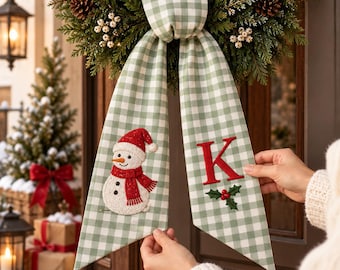May include: A green and white gingham fabric ribbon hanging from a wreath. The left tail features an embroidered snowman with a red hat and scarf. The right tail displays a large red letter K above a small green holly sprig with red berries.