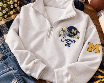 May include: A light gray quarter-zip sweatshirt featuring an embroidered design on the chest with a dark blue football helmet, white daisies, the name Emma, and the number 22. A yellow letter M is embroidered on the left sleeve.