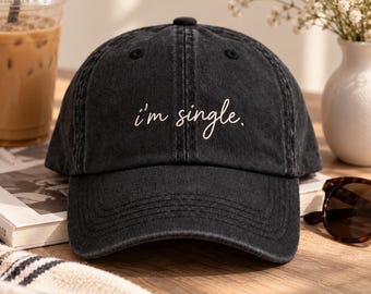 May include: A black baseball cap with a washed-out, distressed texture. The front of the cap features the white embroidered text "i'm single." in a cursive, handwritten-style font. The cap has a structured crown and a curved brim.