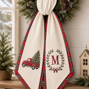 May include: A cream-coloured fabric table runner featuring a red plaid border. One end displays an embroidered red pickup truck carrying a green pine tree, while the other end features a red letter M inside a green wreath with a red bow.