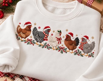 May include: A white crewneck sweatshirt featuring an embroidered design of five chickens in a row. Each chicken wears a red Santa hat, with some wearing red and white striped scarves or bows, surrounded by green pine branches and red berries.