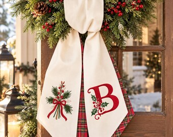 May include: A cream-colored fabric door hanger featuring a festive wreath top with pine needles, pinecones, and red berries. The two hanging tails display an embroidered bouquet of greenery with a red bow and a large red letter B with holly.