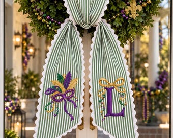May include: A green and white striped fabric door hanger with scalloped edges. The left side features an embroidered purple, green, and gold masquerade mask. The right side displays a large purple letter L topped with a gold bow.
