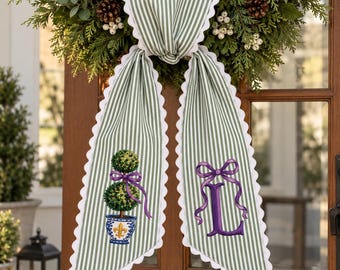 May include: A green and white striped fabric door hanger with scalloped white edges. The left side features an embroidered topiary in a blue and white pot with a purple bow, and the right side features a large purple embroidered letter L.