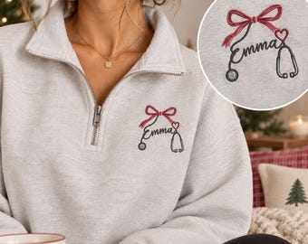 May include: A light gray quarter-zip sweatshirt featuring an embroidered design on the left chest. The embroidery depicts a stethoscope forming the name Emma, accented with a red bow and a small red heart above the stethoscope tubing.