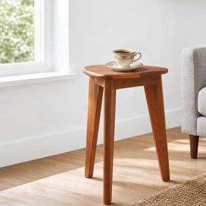 May include: A three-legged wooden side table with a triangular top surface. A ceramic coffee cup with a decorative rim and a matching saucer sits on the table. The table is constructed from a warm-toned wood with a smooth, polished finish.