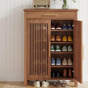 May include: A tall wooden shoe cabinet with a slatted door design and a single top drawer. The cabinet features gold-toned handles and multiple interior shelves filled with various pairs of shoes, including sneakers, loafers, and boots.