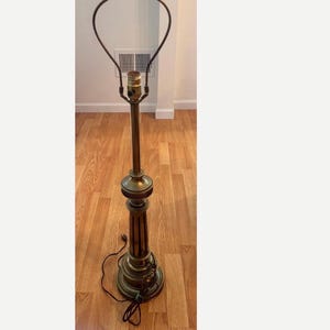 May include: A tall, antique-style brass table lamp base featuring a slender, fluted column, a decorative central bulbous section, and a circular tiered base. The lamp includes a metal harp attachment at the top for a shade and a black electrical cord.